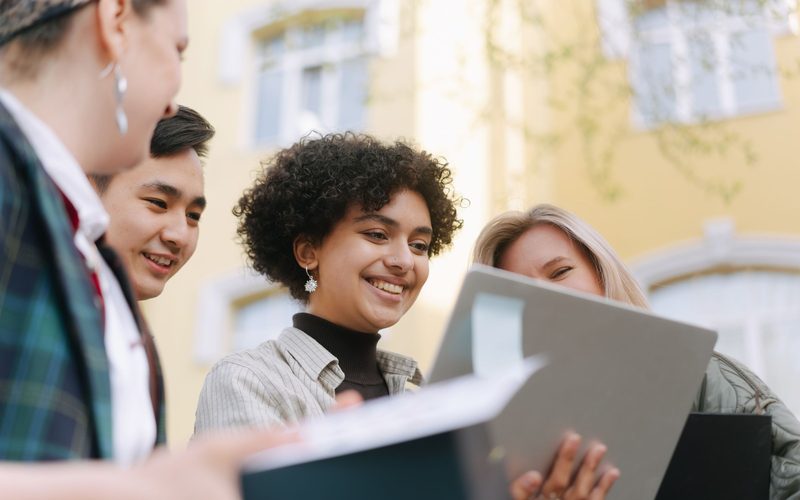 Four students outdoors smiling over a laptop and a sheaf of papers