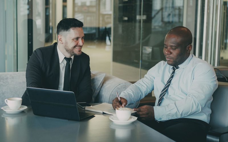 Two people in business dress at a table with a laptop, notes and coffee, one of them signing